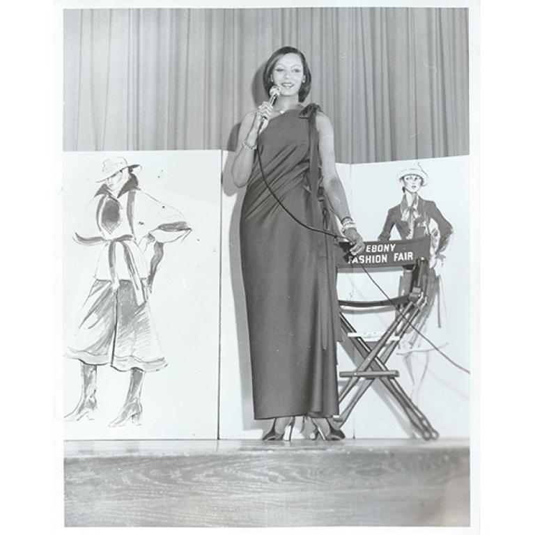 Vintage photo of Audrey Smaltz standing on stage in formal wear and holding microphone.